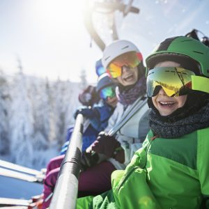 Mother skiing with kids on a sunny winter day. Family is sitting on chairlift cheering at the camera.
Nikon D850